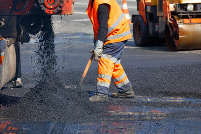 Bild vergrern: A road worker in orange overalls renovates a section of the road with hot asphalt against the background of a road roller in blur. Road repair concept, place for text, copy space.